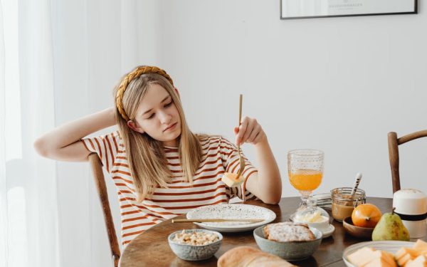Teen girl in striped shirt at breakfast table with fruits, bread, and juice. Casual morning setting.