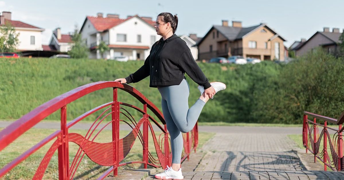 A woman in active wear performs a stretching exercise on a red bridge in a suburban park setting.