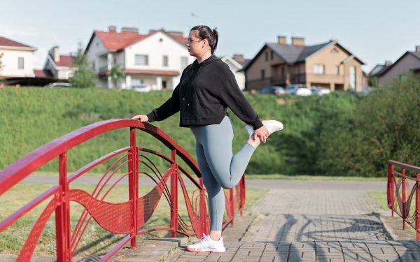 A woman in active wear performs a stretching exercise on a red bridge in a suburban park setting.